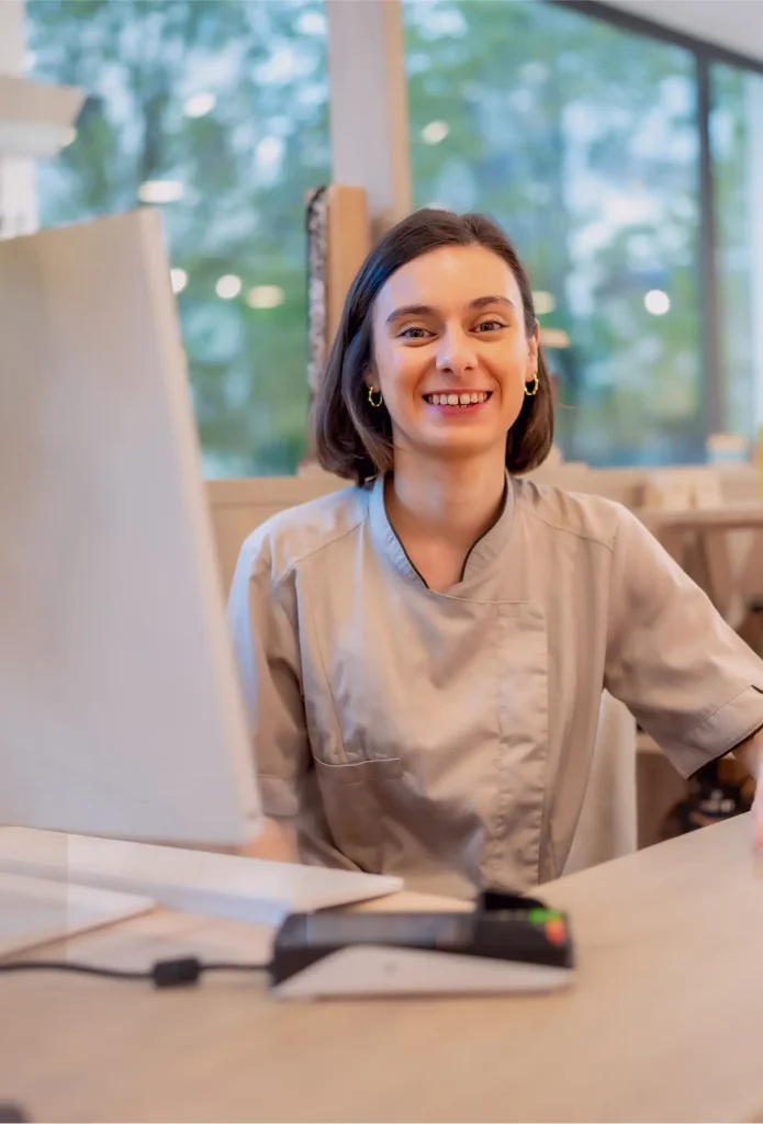 A woman in a gray uniform sits at a desk with a computer, smiling at the camera in a bright, modern office setting.