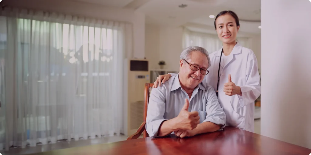An older man sits at a table giving a thumbs up, while a healthcare professional stands beside him, also giving a thumbs up. Both are smiling in a bright room.
