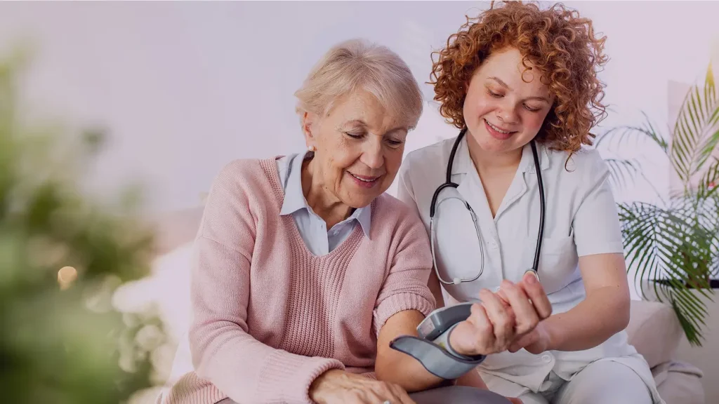 A nurse in a white uniform checks the blood pressure of an older woman in a pink sweater. Both are smiling and sitting indoors.