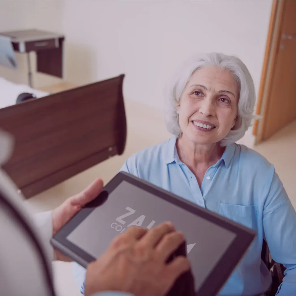 An older woman with gray hair sits in a room, smiling at someone who is holding a tablet in front of her.