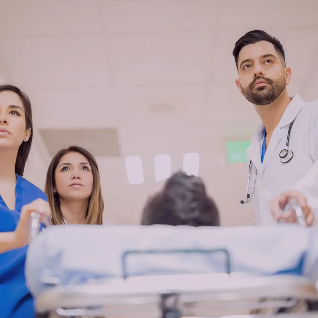 Three medical professionals escort a patient on a stretcher down a hospital hallway, looking ahead with serious expressions.