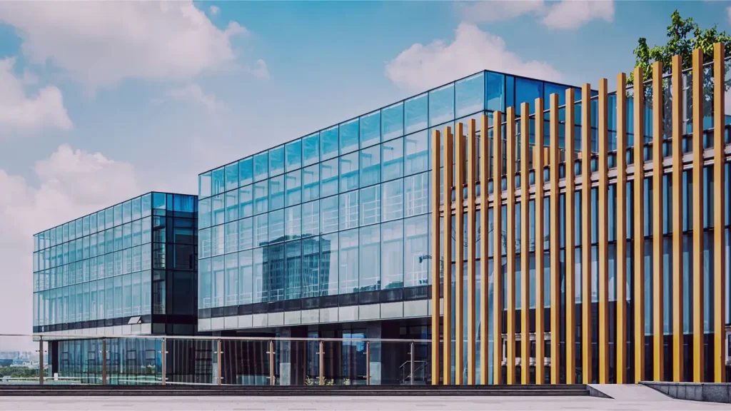Modern glass office building with wooden vertical slats on the right side, set against a blue sky with scattered clouds.